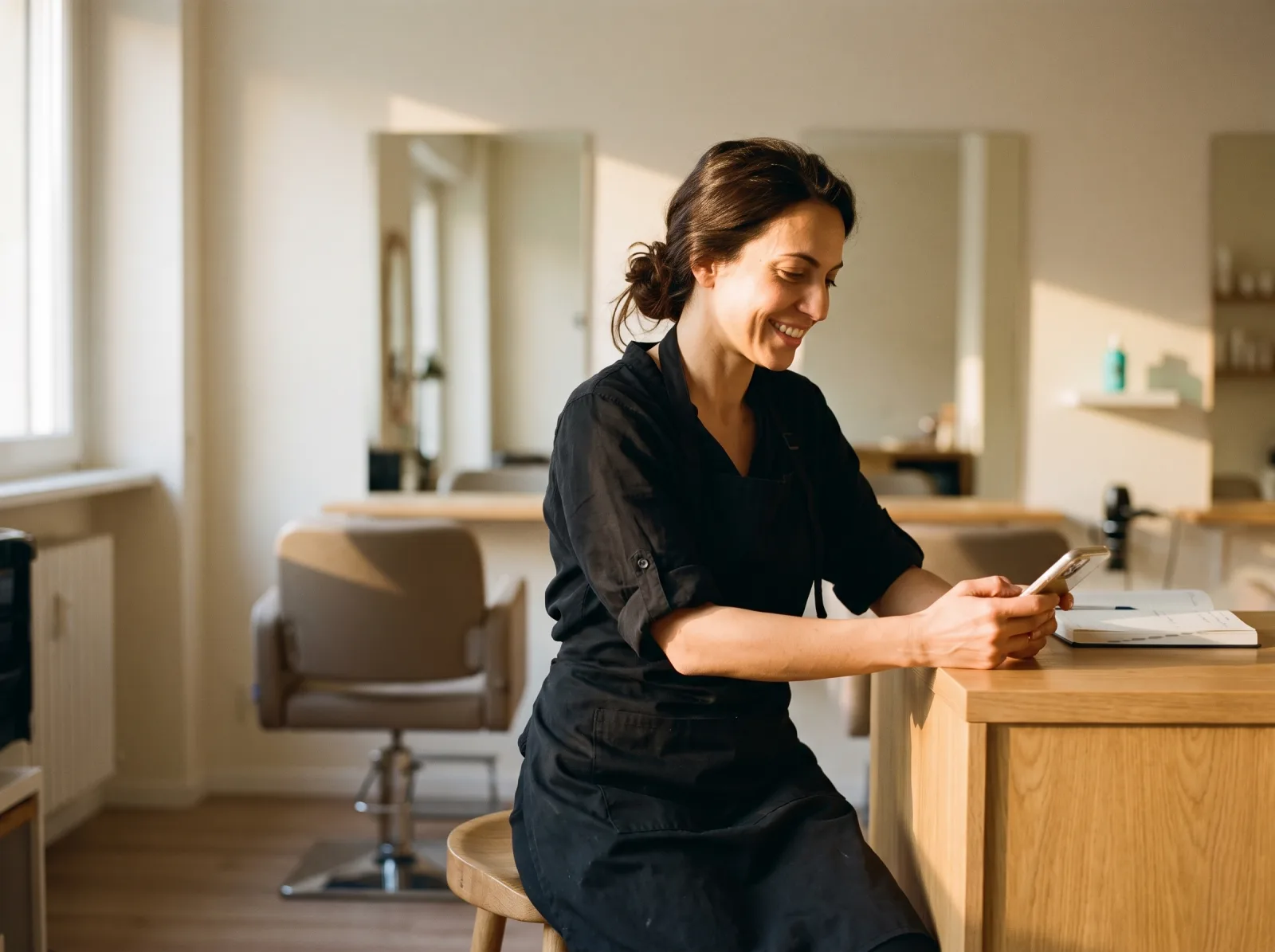 La titolare seduta al bancone a fine giornata, viso illuminato e sorriso, manda dal telefono gli ultimi promemoria mentre entra la luce lunga del pomeriggio